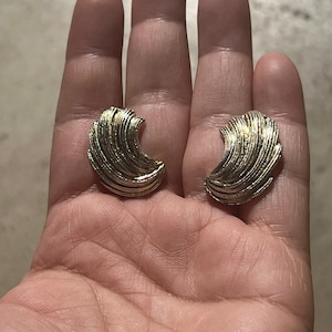 May include: A pair of gold-toned earrings with a crescent moon shape. The earrings are made of a textured metal and have a slightly hammered finish.