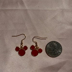 May include: A pair of gold-tone earrings with red Mickey Mouse-shaped charms. The earrings are about the size of a US quarter.