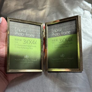 May include: A gold-colored, hinged photo frame with two sections. Each section displays the text "Empress Photo Frame" and dimensions of 3 1/4 x 4 1/4 inches. The frame is from Acme Frame Products Inc., Chicago.