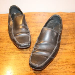 Puede incluir: Un par de mocasines de cuero negro con un dise&ntilde;o cosido. Los mocasines est&aacute;n sobre una superficie de madera.