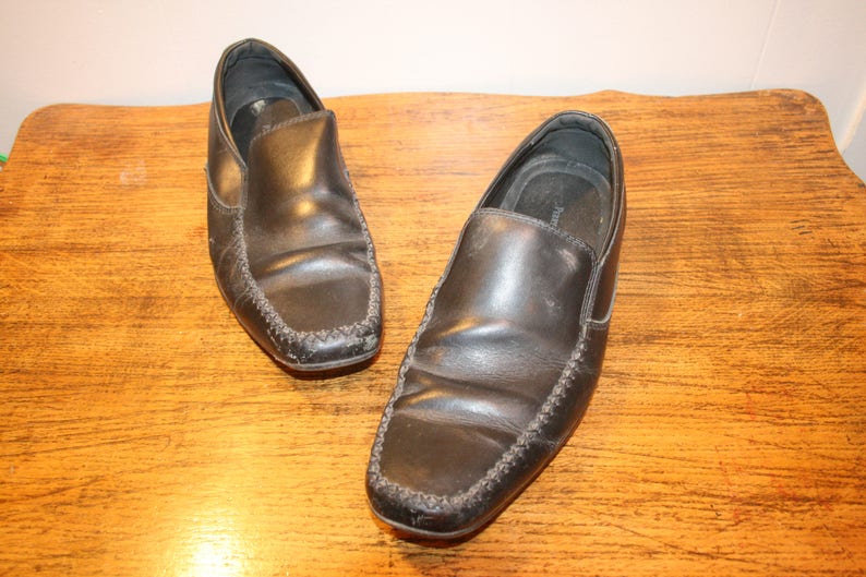 Puede incluir: Un par de mocasines de cuero negro con un dise&ntilde;o cosido. Los mocasines est&aacute;n sobre una superficie de madera.