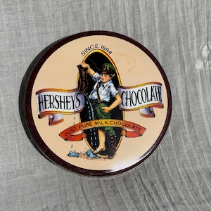 May include: Round Hershey's Chocolate tin with a vintage illustration. The image features a young boy pouring chocolate from a boot. Text includes "Hershey's Chocolate" and "Just Pure Milk Chocolate" with "Since 1894".