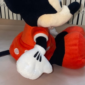 Op de afbeelding: Een pluche speelgoed van Minnie Mouse die een rode jurk met witte stippen draagt. Ze heeft zwarte oren en een zwarte strik. Haar ogen zijn gesloten en ze glimlacht.