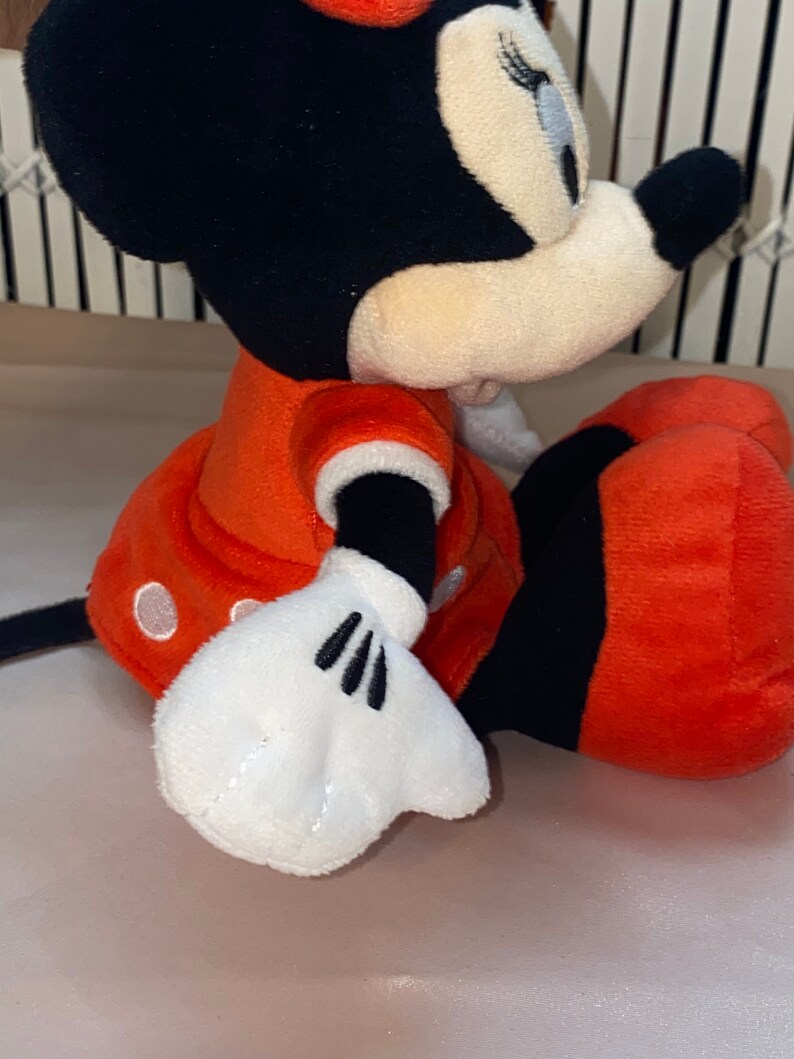 Op de afbeelding: Een pluche speelgoed van Minnie Mouse die een rode jurk met witte stippen draagt. Ze heeft zwarte oren en een zwarte strik. Haar ogen zijn gesloten en ze glimlacht.