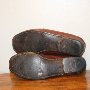 Op de afbeelding: Een paar bruine leren loafers met versleten leren zolen. De loafers liggen op elkaar.