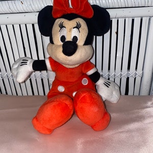 Op de afbeelding: Een pluche speelgoed van Minnie Mouse die een rode jurk draagt met witte stippen en een rode strik. Ze heeft zwarte oren en een zwarte neus.