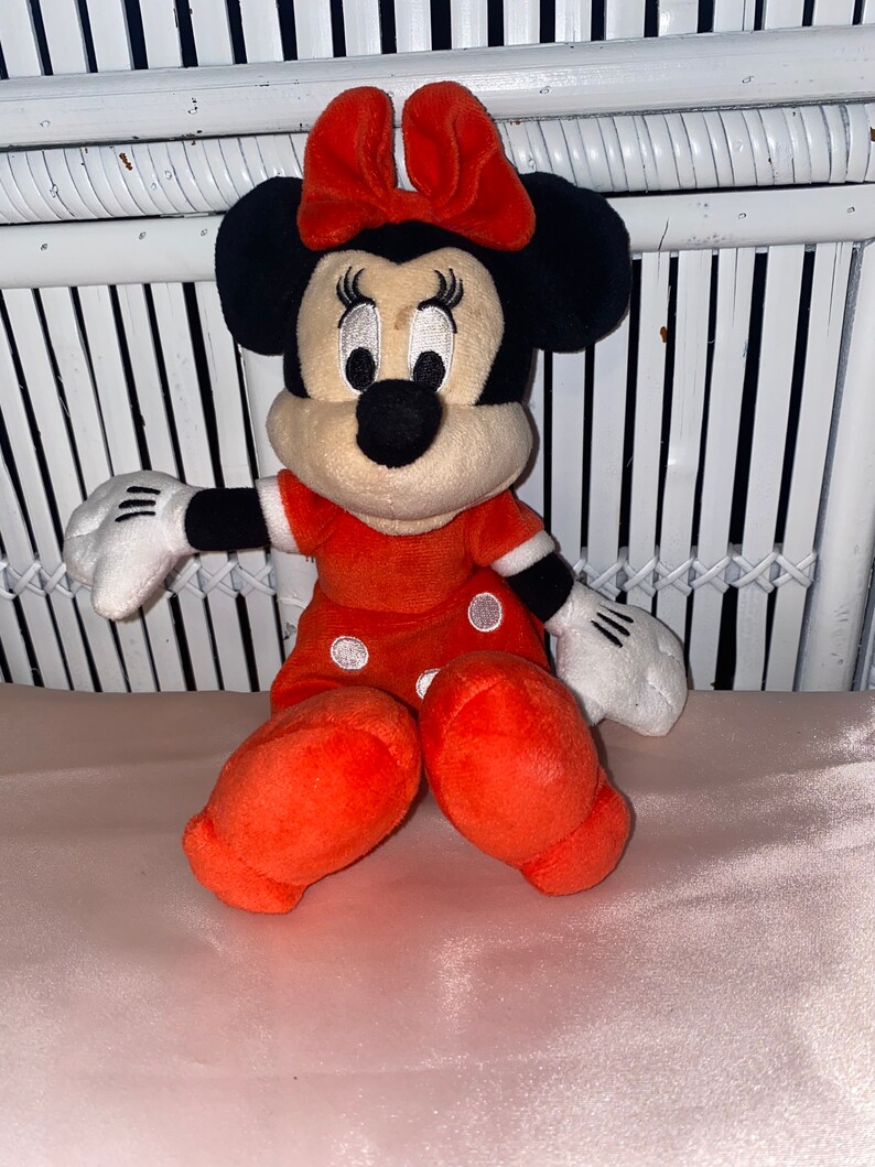 Op de afbeelding: Een pluche speelgoed van Minnie Mouse die een rode jurk draagt met witte stippen en een rode strik. Ze heeft zwarte oren en een zwarte neus.