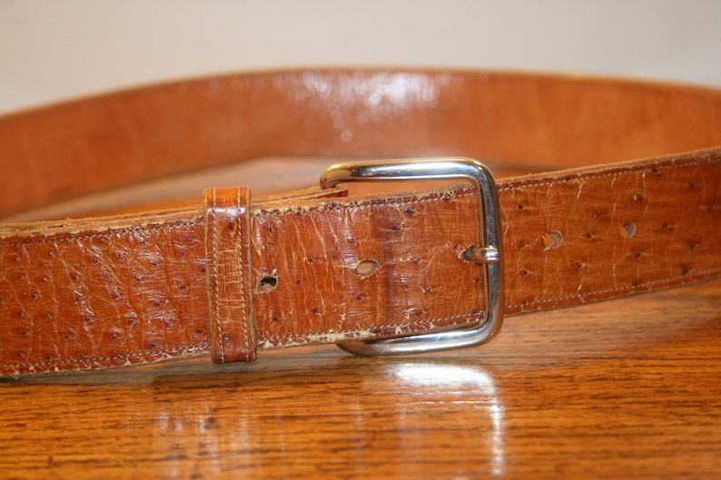 Op de afbeelding: Een bruine leren riem met een zilveren gesp. De riem heeft een textuur, vergelijkbaar met die van een struisvogel.