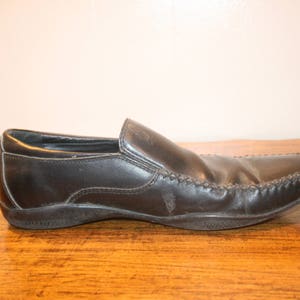 Puede incluir: Un mocas&iacute;n de cuero negro con un detalle cosido alrededor de la puntera. El zapato tiene un dise&ntilde;o de deslizamiento y una puntera ligeramente redondeada.