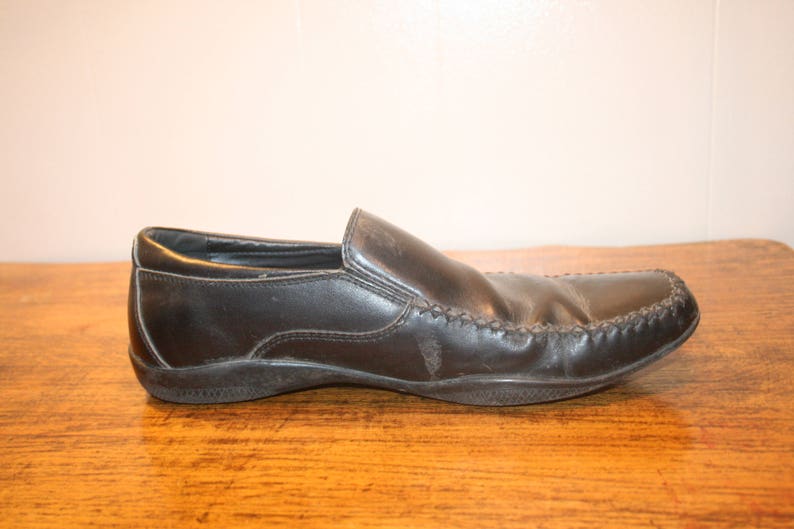Puede incluir: Un mocas&iacute;n de cuero negro con un detalle cosido alrededor de la puntera. El zapato tiene un dise&ntilde;o de deslizamiento y una puntera ligeramente redondeada.