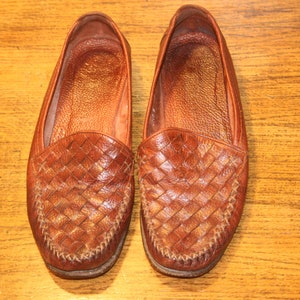 Op de afbeelding: Een paar bruine leren loafers met een geweven patroon. De loafers hebben een spitse neus en een gestikte naad rond de rand.