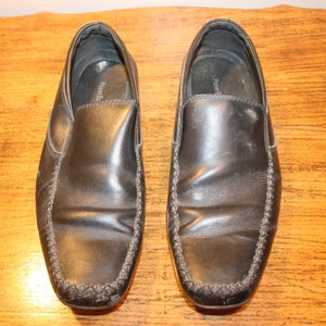 Puede incluir: Un par de zapatos negros de cuero sin cordones con un detalle cosido alrededor del borde.