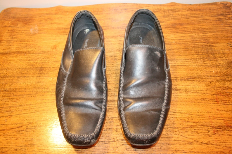 Puede incluir: Un par de zapatos negros de cuero sin cordones con un detalle cosido alrededor del borde.