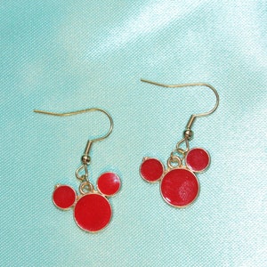 May include: A pair of gold dangle earrings with red circle charms. The charms are arranged in a Mickey Mouse shape.