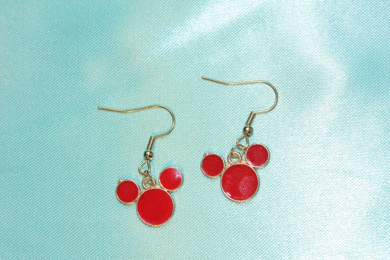 May include: A pair of gold dangle earrings with red circle charms. The charms are arranged in a Mickey Mouse shape.
