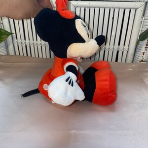 Op de afbeelding: Een pluche rood en zwart Minnie Mouse speelgoed met witte handschoenen en een rode strik. Het speelgoed zit op een wit oppervlak.