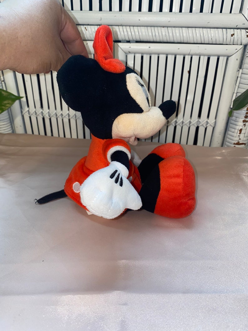 Op de afbeelding: Een pluche rood en zwart Minnie Mouse speelgoed met witte handschoenen en een rode strik. Het speelgoed zit op een wit oppervlak.