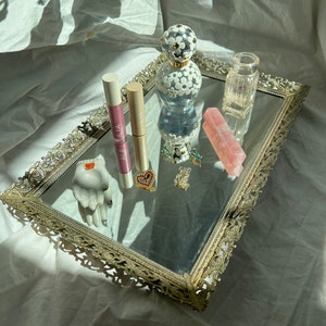 May include: A silver tray with an ornate design, featuring a clear glass bottle with a white floral design, two pink lip glosses, a pink crystal, a small clear glass jar, and two small gold jewellery pieces.