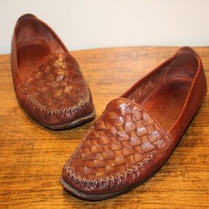 Op de afbeelding: Een paar bruine leren geweven loafers. De loafers hebben een spitse neus en een gestikte naad rond de rand.