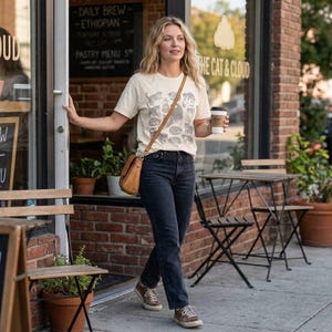Op de afbeelding: Een vrouw draagt een crèmekleurig t-shirt met een kattenafbeelding, een donkerblauwe jeans en een bruine leren crossbodytas. Ze houdt een koffiekopje vast en staat voor een café met de tekst "THE CAT & CLOUD".