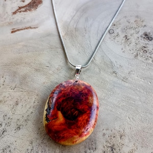 May include: A handcrafted pendant necklace featuring a large, oval-shaped wooden pendant with a vibrant, swirling pattern of red, orange, and brown hues. The pendant is suspended from a delicate silver chain.