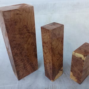 Op de afbeelding: Drie onafgewerkte houten blokken met een rijke, roodbruine kleur en unieke nerfpatronen. De blokken variëren in grootte en vorm, met één lang en rechthoekig, één vierkant en één korter en rechthoekig. Het hout heeft een natuurlijke, organische uitstraling.