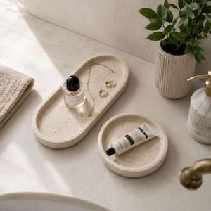 May include: An oval-shaped beige speckled tray holding a small glass perfume bottle with a black cap, a gold chain necklace, and two gold hoop earrings, alongside a circular beige speckled tray containing a white tube of hand cream.
