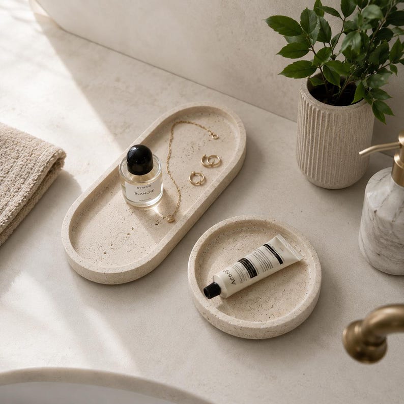 May include: An oval-shaped beige speckled tray holding a small glass perfume bottle with a black cap, a gold chain necklace, and two gold hoop earrings, alongside a circular beige speckled tray containing a white tube of hand cream.