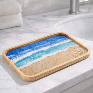 May include: A rectangular bamboo tray with a resin design depicting a beach scene. The tray features a light brown wooden frame, with a blue and white ocean design and a sandy beach. The tray is on a white marble surface.