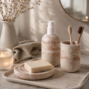 Puede incluir: Un juego de ba&ntilde;o de piedra beige que incluye un dispensador de jab&oacute;n, un soporte para cepillos de dientes, un jabonera y una bandeja. Los art&iacute;culos tienen un acabado texturizado con un patr&oacute;n de rayas. Una pastilla de jab&oacute;n con la palabra "meraki" est&aacute; en la jabonera. Dos cepillos de dientes est&aacute;n en el soporte.