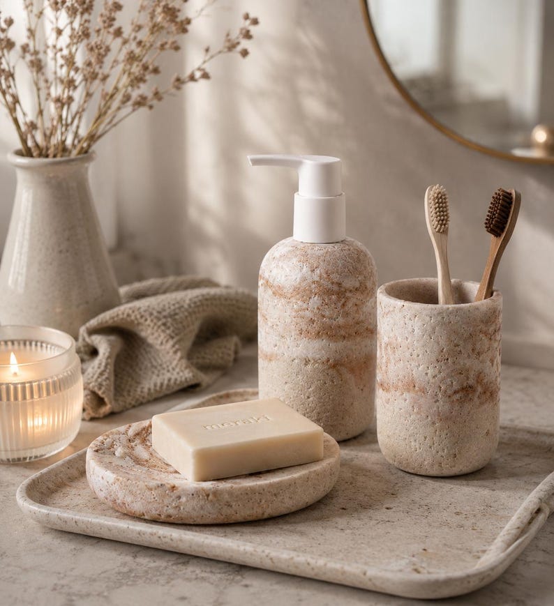 Puede incluir: Un juego de ba&ntilde;o de piedra beige que incluye un dispensador de jab&oacute;n, un soporte para cepillos de dientes, un jabonera y una bandeja. Los art&iacute;culos tienen un acabado texturizado con un patr&oacute;n de rayas. Una pastilla de jab&oacute;n con la palabra "meraki" est&aacute; en la jabonera. Dos cepillos de dientes est&aacute;n en el soporte.