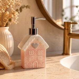 Könnte beinhalten: Ein rosa Keramik-Seifenspender in Form eines Hauses mit weißem Dach und silberner Pumpe. Das Haus hat ein Herz und Fenster. Der Spender steht auf einer Badezimmer-Arbeitsplatte.