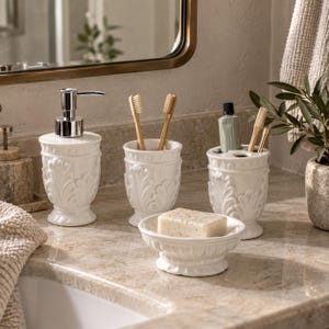 May include: A white ceramic bathroom accessory set featuring a soap dispenser with a silver pump, two toothbrush holders, and a soap dish holding a bar of soap. Each piece is decorated with a raised, embossed leaf pattern around the base.