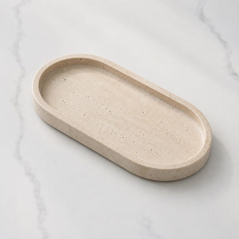 May include: An oval-shaped tray with a light beige, porous, stone-like texture. The tray features a smooth, raised rim and a flat, recessed base with visible natural pitting and variations in the surface finish throughout the entire object.