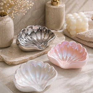 May include: Three seashell-shaped dishes in silver, pink, and white. The dishes have a pearlescent finish and are arranged on a neutral surface. A soap dispenser, candle, and vase are in the background.