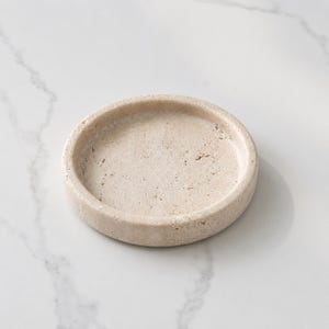May include: A circular beige stone tray with a porous, textured surface and a raised rim. The object features natural variations in color and small indentations throughout the material, creating a distinct, organic appearance for this small dish.