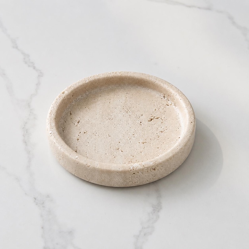 May include: A circular beige stone tray with a porous, textured surface and a raised rim. The object features natural variations in color and small indentations throughout the material, creating a distinct, organic appearance for this small dish.