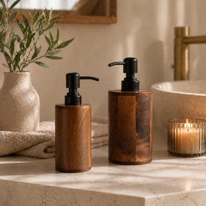 May include: Two wooden soap dispensers with black pumps on a countertop. The dispensers have a cylindrical shape and a natural wood grain finish. A lit candle in a glass holder and a vase with greenery are in the background, suggesting a bathroom setting.