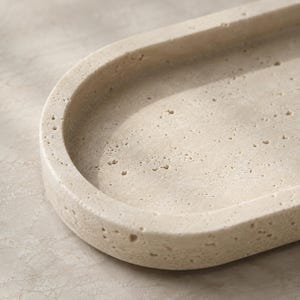 May include: An oval-shaped tray crafted from beige travertine stone, featuring a natural porous texture with small, irregular indentations across its surface. The tray has a smooth, raised rim and a recessed center, showcasing the organic, earthy tones.