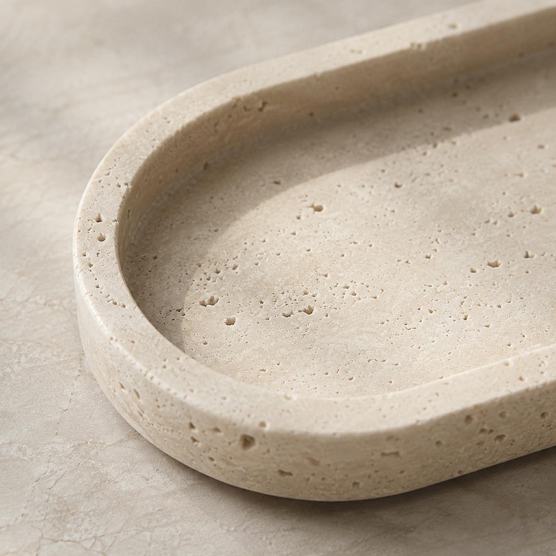 May include: An oval-shaped tray crafted from beige travertine stone, featuring a natural porous texture with small, irregular indentations across its surface. The tray has a smooth, raised rim and a recessed center, showcasing the organic, earthy tones.
