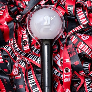 May include: A black and silver light stick with a white orb, featuring a logo, rests amidst a sea of red and black lanyards. The lanyards display the words "WORLD TOUR" and various names in white lettering.