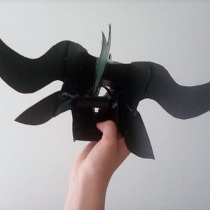 May include: A black, handmade headpiece with curved horns and pointed ear-like shapes. The headpiece has a central structure with green accents, possibly leaves or feathers. The design suggests a fantasy or cosplay theme.