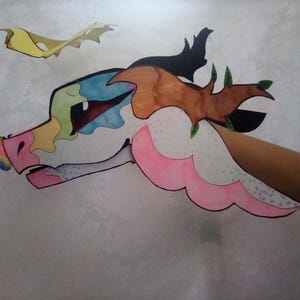 May include: A colorful, handmade paper dragon head with a rainbow horn, pink accents, and a brown horn. The dragon is held by a person's arm. Two yellow paper cutouts are in the background.