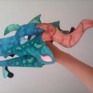 May include: A handmade dragon head puppet in shades of teal, blue, and green, with brown horns and pink accents. The puppet is held up by a person's arm, showcasing the detailed, colorful design.