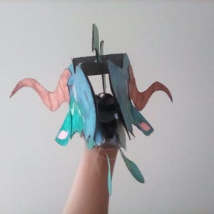 May include: A handmade papercraft dragon head with brown horns, blue wings, and green accents. The dragon head is constructed from various colored papers and is held up by a hand.
