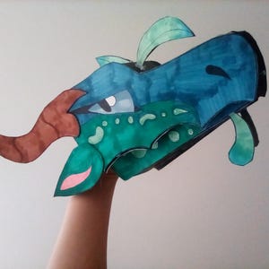 May include: A handmade, colorful dragon mask. The mask features shades of blue, green, and brown, with a pink accent. The dragon's head is detailed with eyes, horns, and leaf-like features, creating a whimsical and imaginative design.