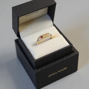 Op de afbeelding: Een gouden ring met een kleine, vierkante rode edelsteen in een geometrisch ontwerp, gepresenteerd in een zwarte sieradendoos met een wit interieur. De doos is gemarkeerd met "JEWELS TEASURE".