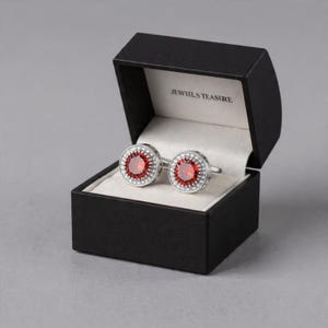 May include: Silver cufflinks with a central red gemstone and a halo of small white stones, presented in an open black box. The box interior is white, and the text "JEWILS TEASURE" is visible.