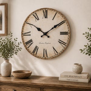 May include: A round wall clock with a distressed beige face, black Roman numerals, and black metal hands. The clock face features the text London, England in a script font. The clock has a thin, dark-toned frame and is mounted on a wall.