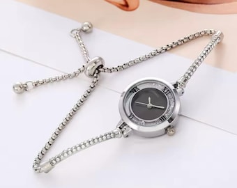 Könnte beinhalten: Eine silberfarbene analoge Armbanduhr mit einem runden schwarzen Zifferblatt und römischen Ziffern auf den Positionen zwölf, drei, sechs und neun Uhr. Die Lünette ist mit klaren Kristallen besetzt und das Armband ist eine flexible, verstellbare Kette.